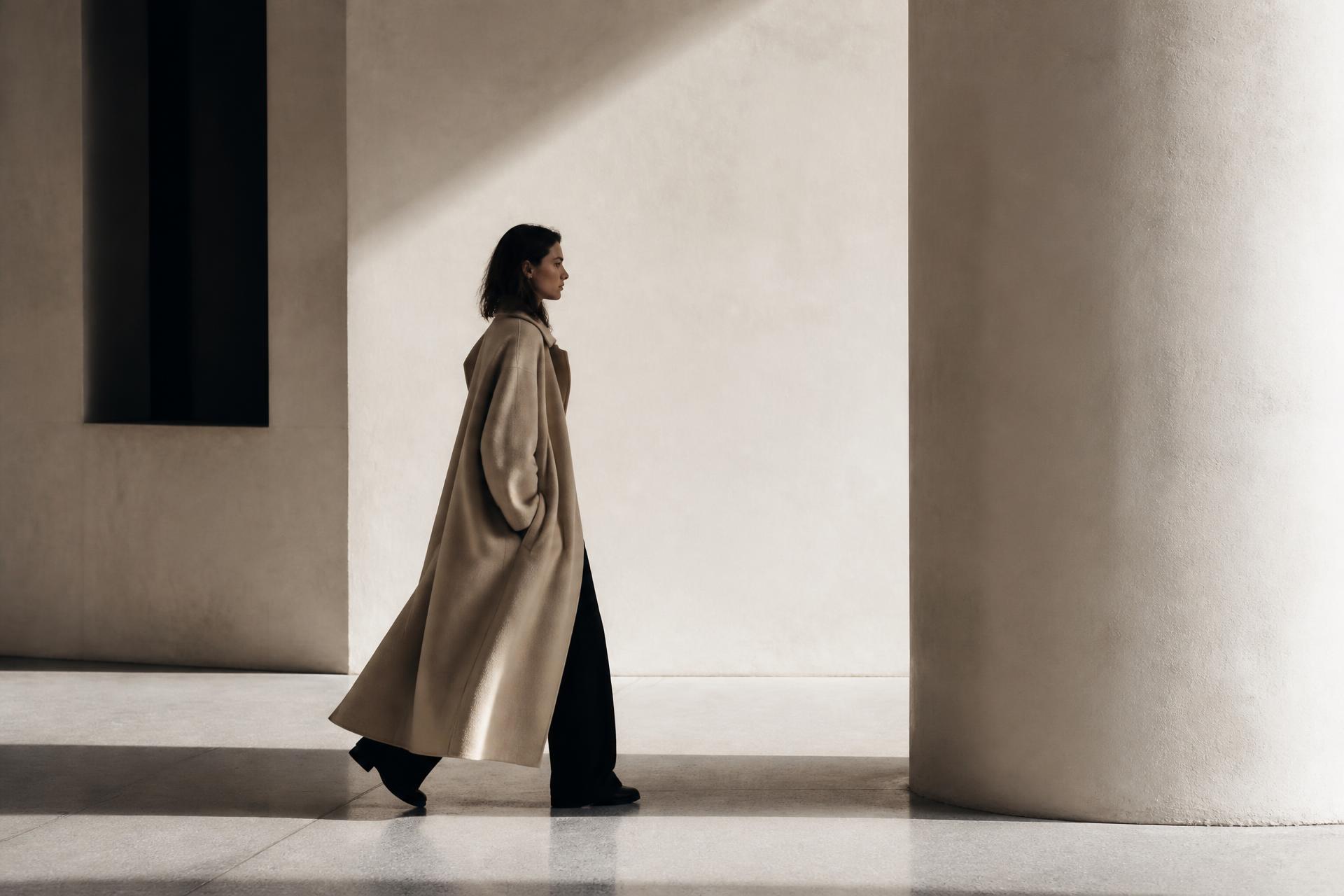 Figure in an oversized beige coat crossing a sunlit minimal gallery space